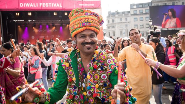 Diwali im Trafalgar Square. Bild mit Genehmigung von the Greater London Authority. Ein Mann in traditioneller Kleidung mit Blumengirlanden um den Hals auf dem Diwali Festival, während andere Schausteller im Hintergrund tanzen.