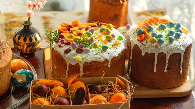 Ein Tablett mit orangefarbenen Eiern im Stroh und zwei große Osterkuchen mit bunten Eiern und weißem Zuckerguss. 
