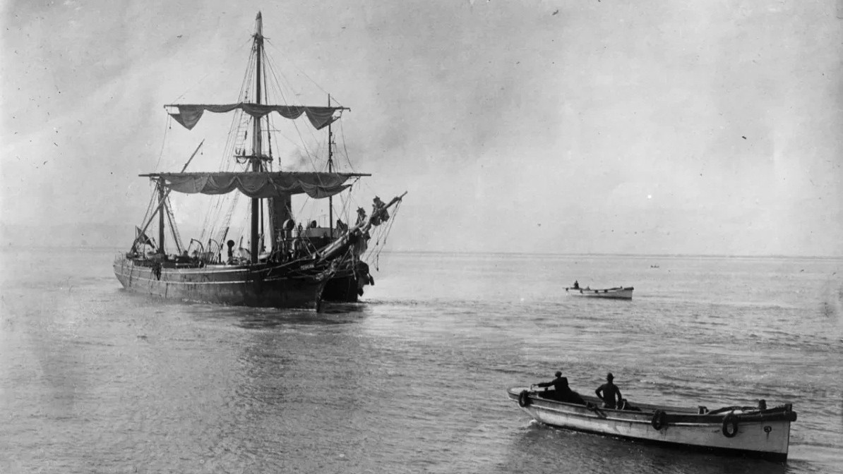 A black-and-white photograph of Ferreira ship with its rigging on the water and a smaller boat in the foreground.