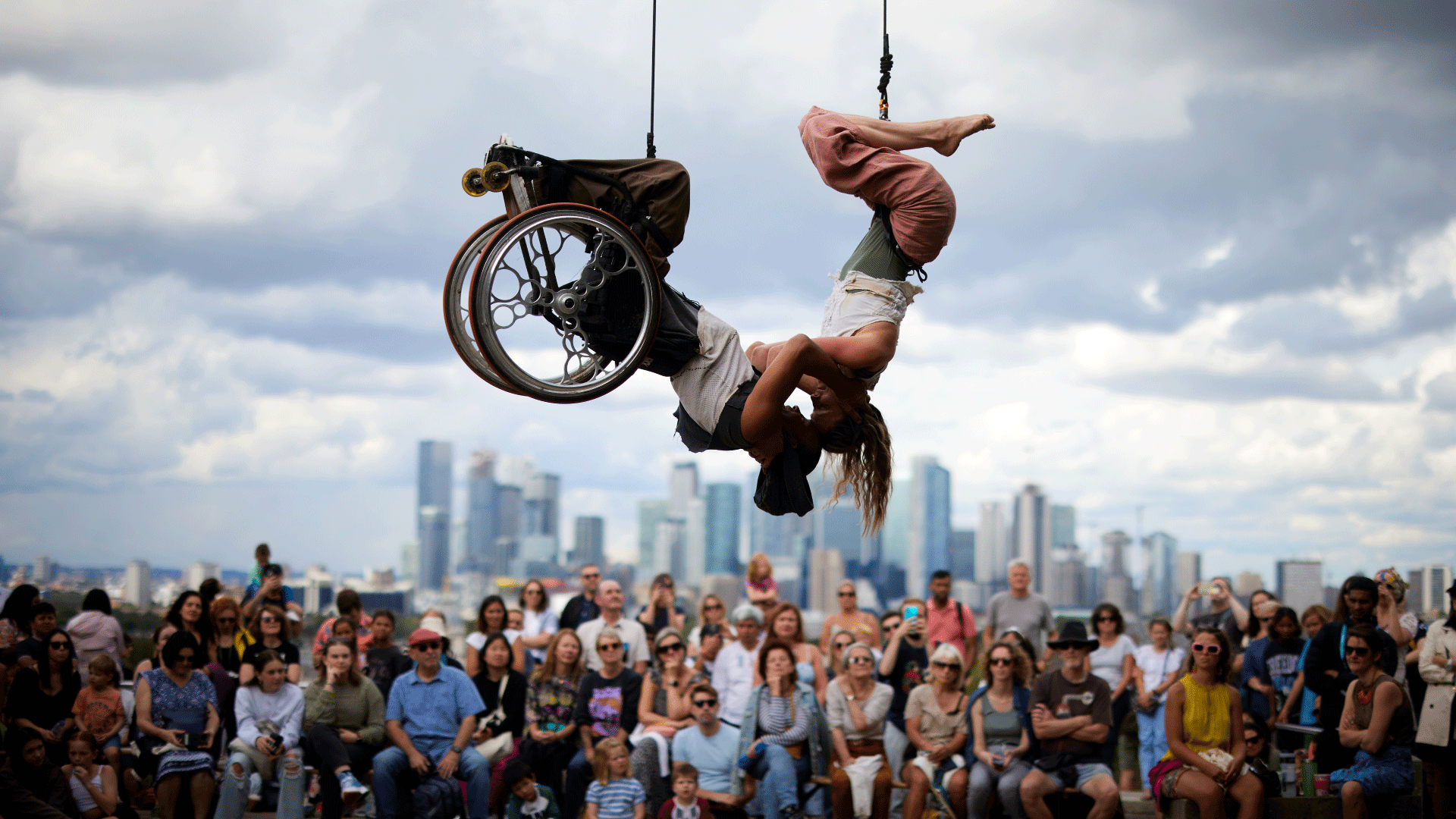 Two performers hang from ropes during a performance in front of crowds with the City of London in the background