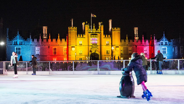 Une jeune fille patinant avec un pingouin sur la patinoire de nuit devant le palais de Hampton Court éclairé de lumières rouges, jaunes et bleues.