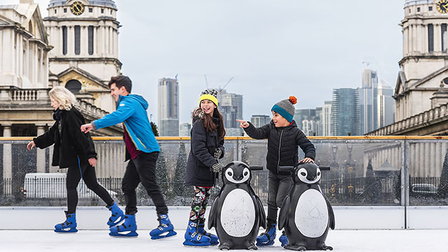 Two smiling kids stand next to penguin-shaped skating aids on an outdoor rink at The Queen's House Ice Rink in Greenwich. 