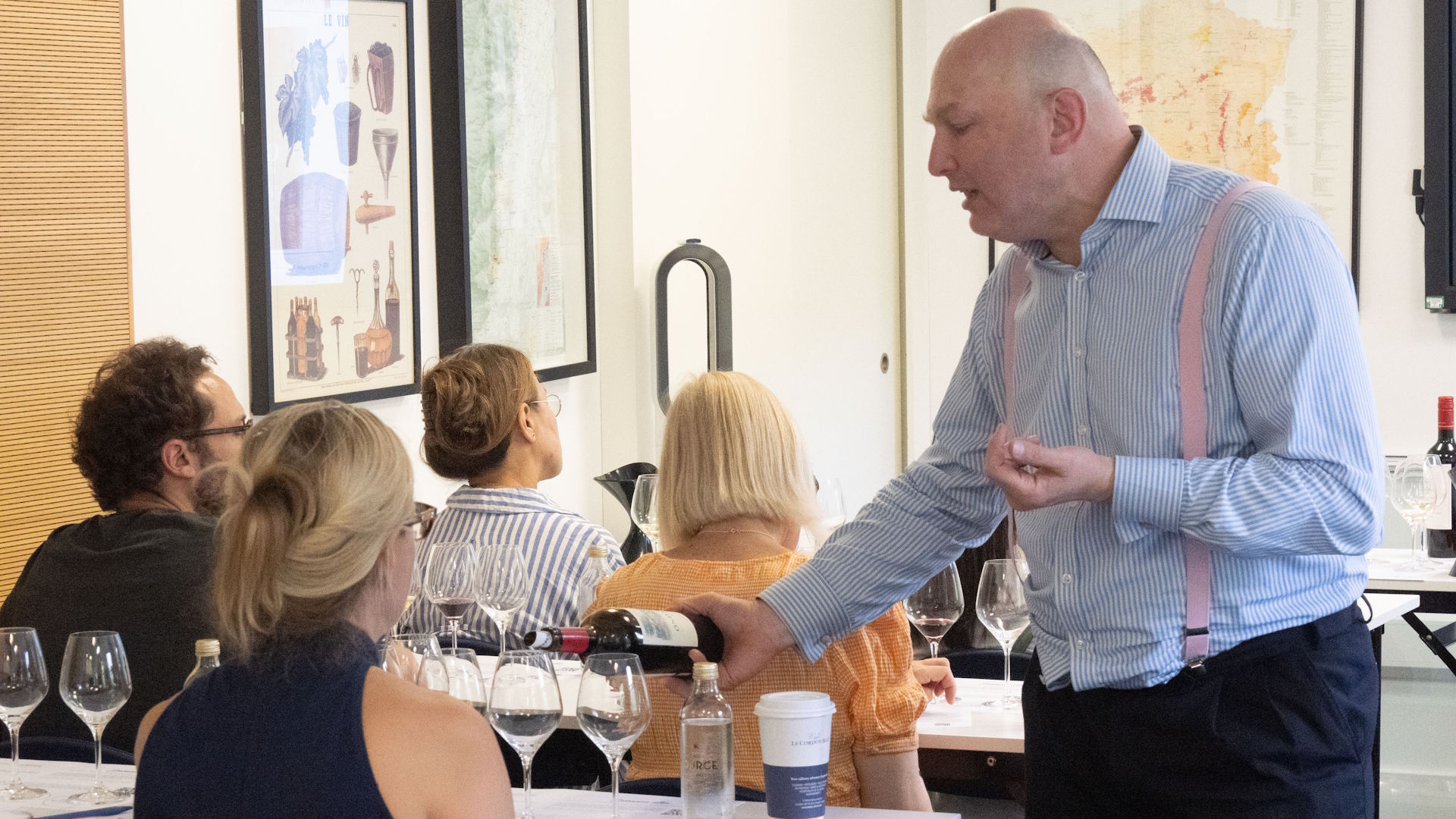 A man pours a glass of wine for a woman during a wine tasting in a classroom, with various empty glasses on each table.