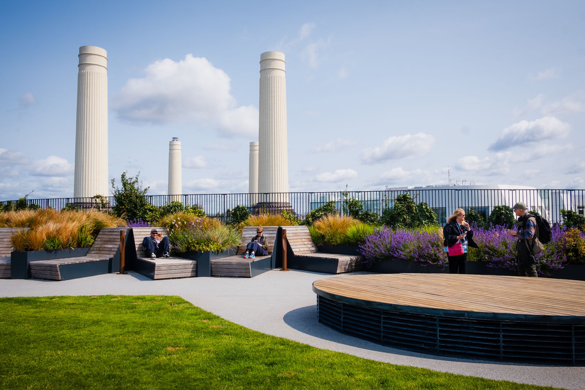 A view of Battersea Roof Gardens, showing people relaxing on benches, borders full of colourful plants and the iconic chimney of Battersea Power Station in the background.