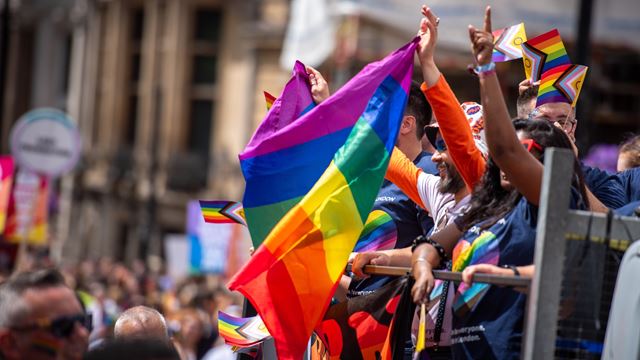 People holding up a multicoloured LGBTQ+ flag aboard a bus parading through the streets of central London for Pride on a sunny day.