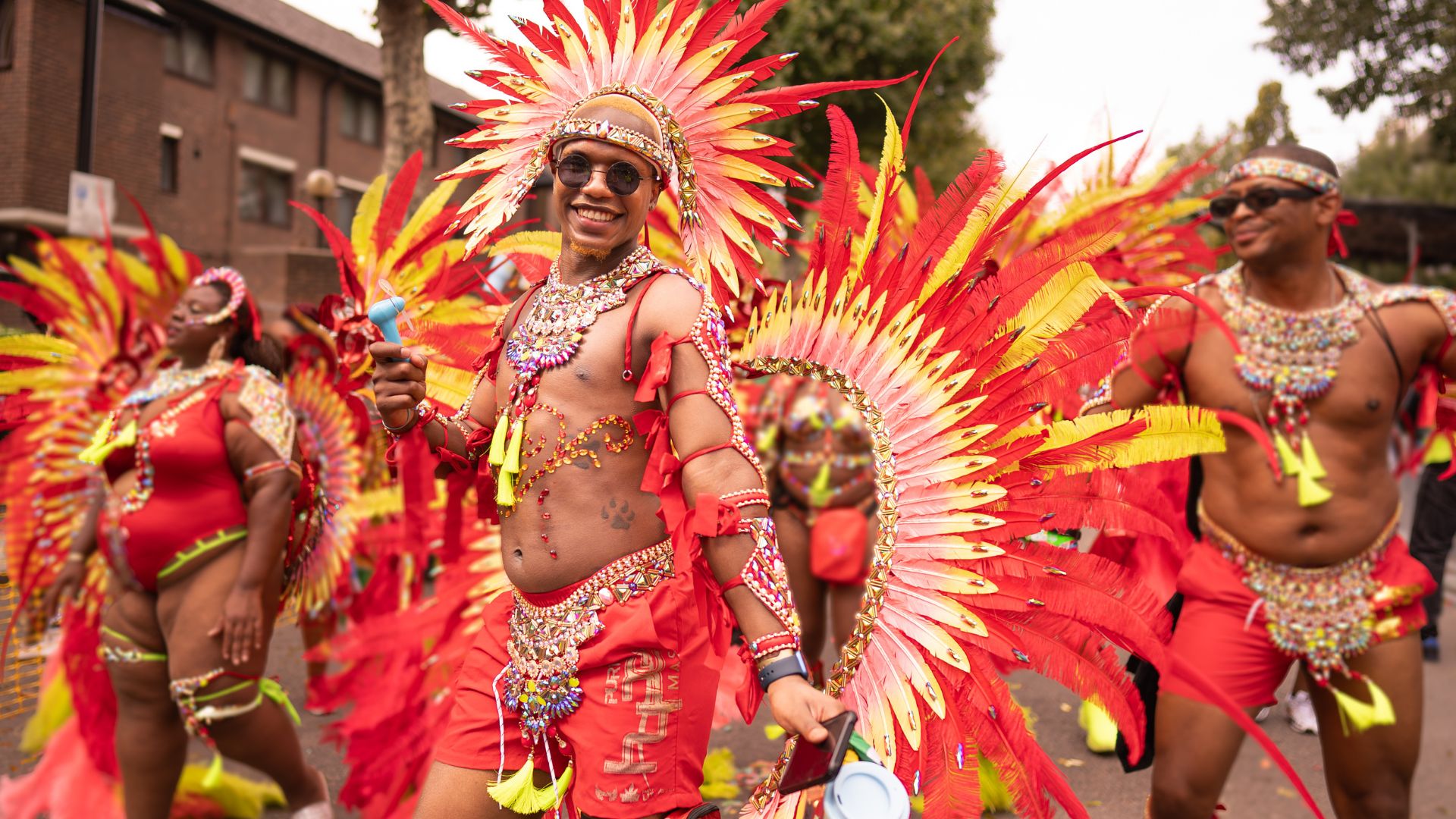 A carnival performer smiles while wearing an elaborate red, orange and gold feathered costume during Notting Hill Carnival. Image credit: London & Partners/Peter Cohen.