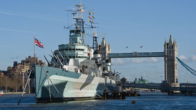 A battleship sits atop the water with a bridge and blue sky in the background. 