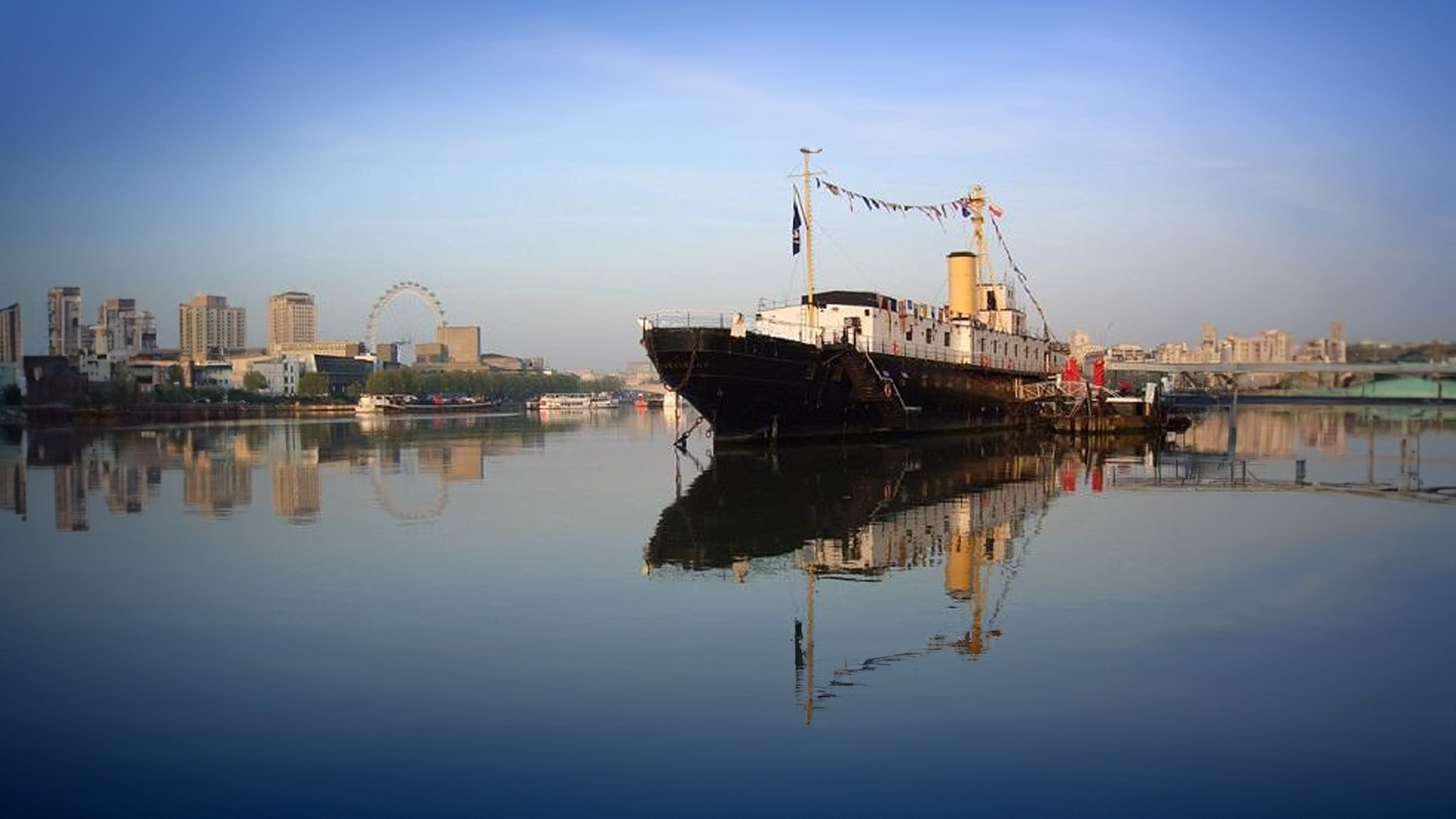 An old boat sits on top of a river with a blue sky and city backdrop