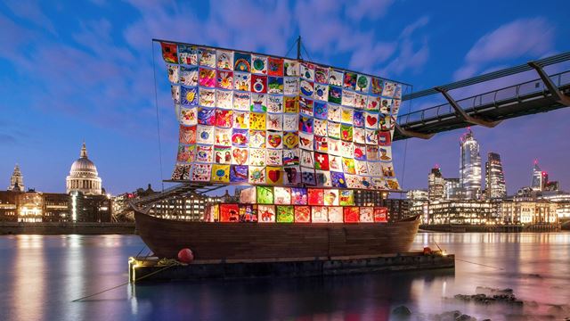 The colourful patchwork sails of The Ship of Tolerance lit up on the Thames at night, with the Millennium Bridge and St Paul's Cathedral in the background. 