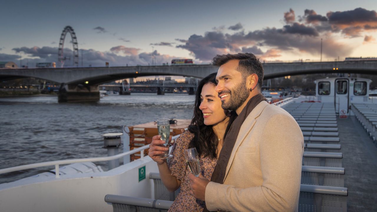 Two people stand on the deck of a river cruise boat drinking champagne.