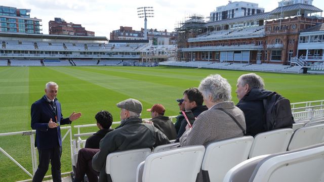 A group of visitors sitting the stands at Lord's Cricket Ground while a tour guide talks.