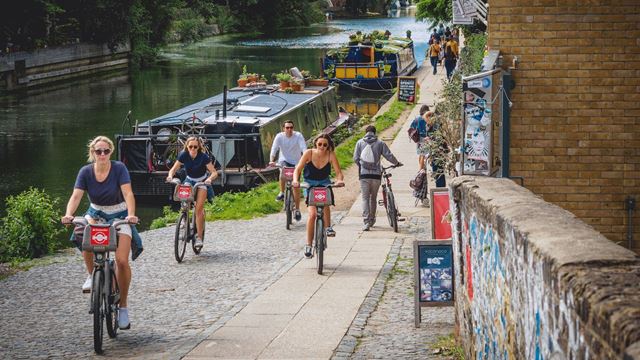People cycling along a canal towpath beside moored narrowboats in London, with pedestrians walking along the waterside.