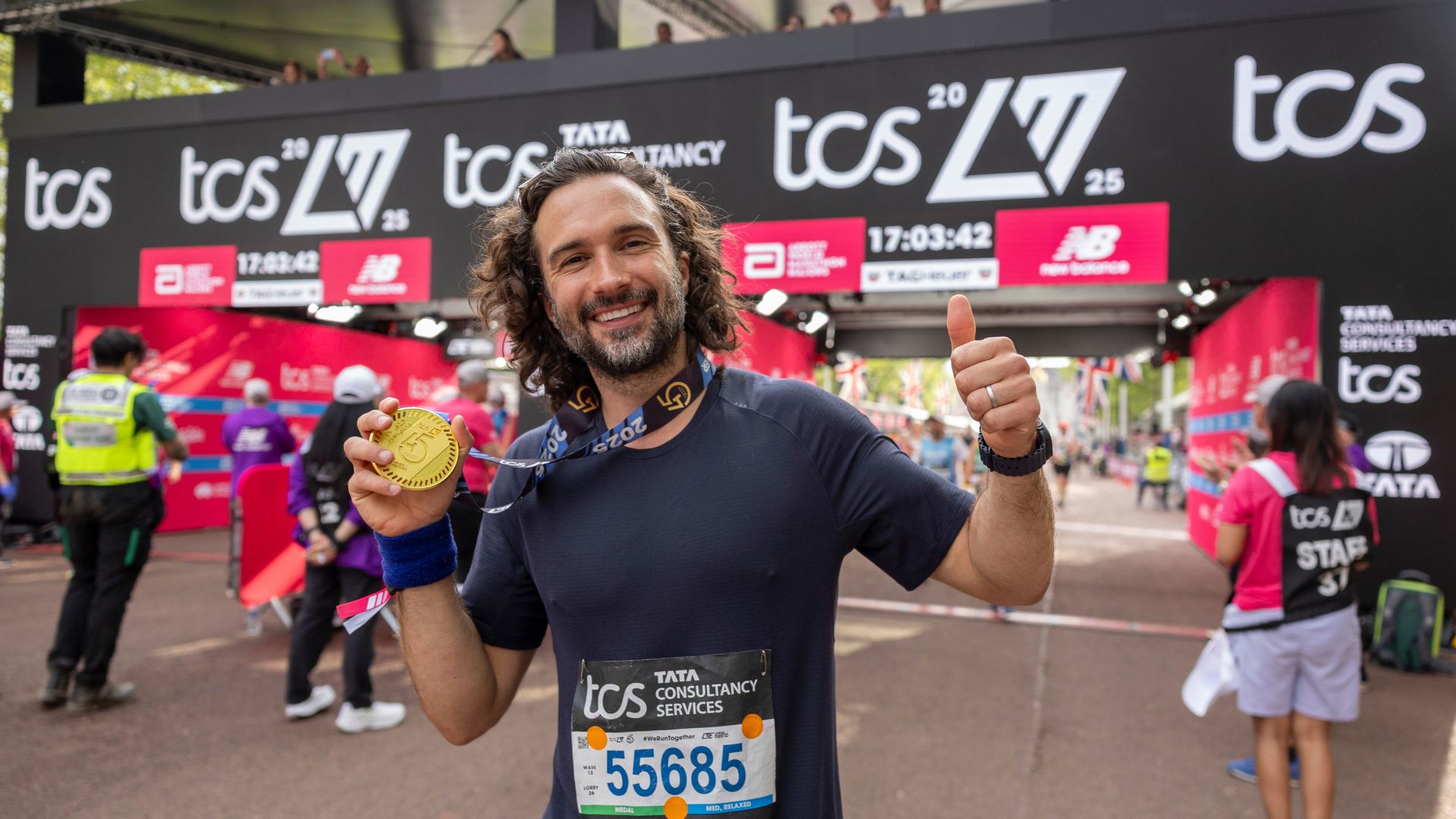 Joe Wicks smiling at the London Marathon finish line, holding up his medal and giving a thumbs up, wearing a race bib with event signage and other runners in the background.