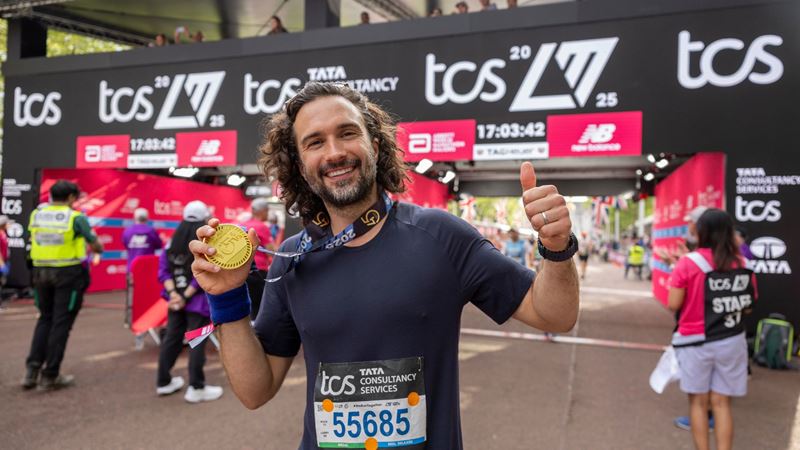 Joe Wicks smiling at the London Marathon finish line, holding up his medal and giving a thumbs up, wearing a race bib with event signage and other runners in the background.
