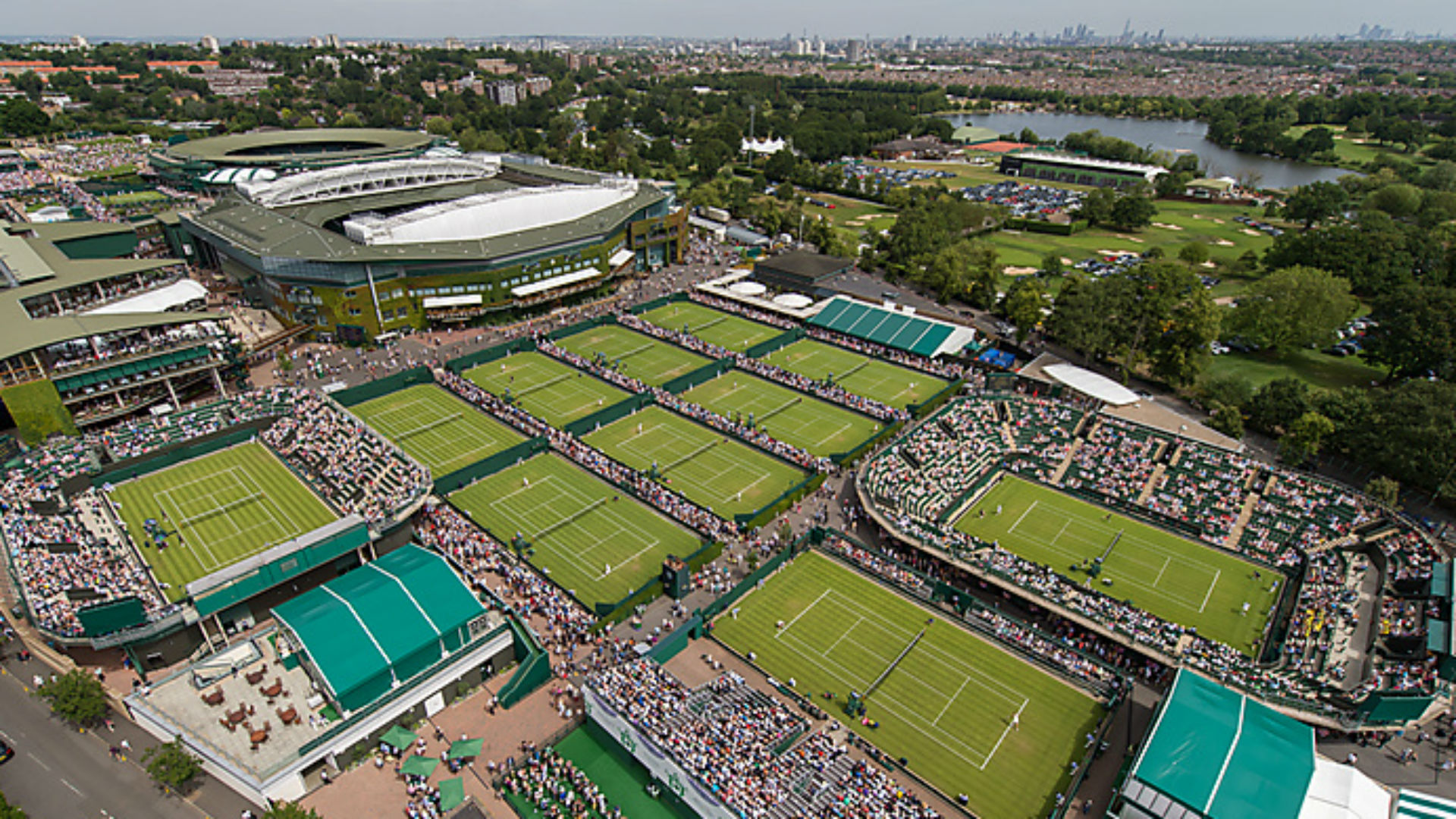 An aerial view of the grounds overlooking Wimbledon on day one of The Championships 2015 at The All England Lawn Tennis Club. Image courtesy of: AELTC/Bob Martin. An aerial view of the tennis courts overlooking Wimbledon on day one of The Championships 2015 at The All England Lawn Tennis Club with London's panorama in the background