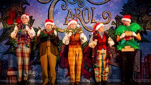 Five actors on stage in festive costumes with Santa's hats and Carol written in big letters on a screen behind them.