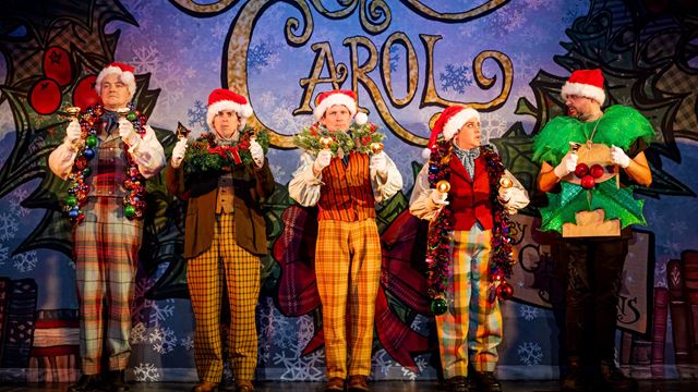 Five actors on stage in festive costumes with Santa's hats and Carol written in big letters on a screen behind them.
