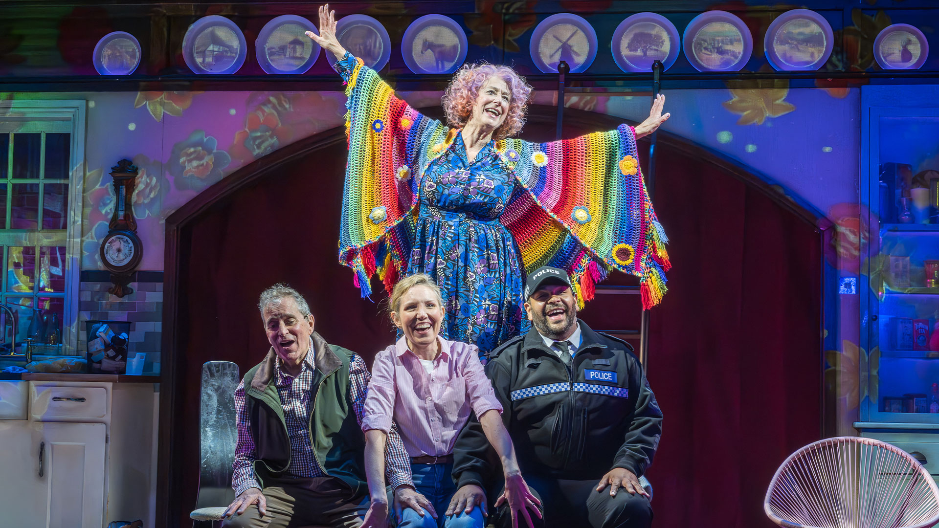 Wearing a blue dress and a rainbow-coloured knitted poncho, Maureen Lipman stands on a table, raising her hands in the air. The other cast members are seated and smiling at the audience.