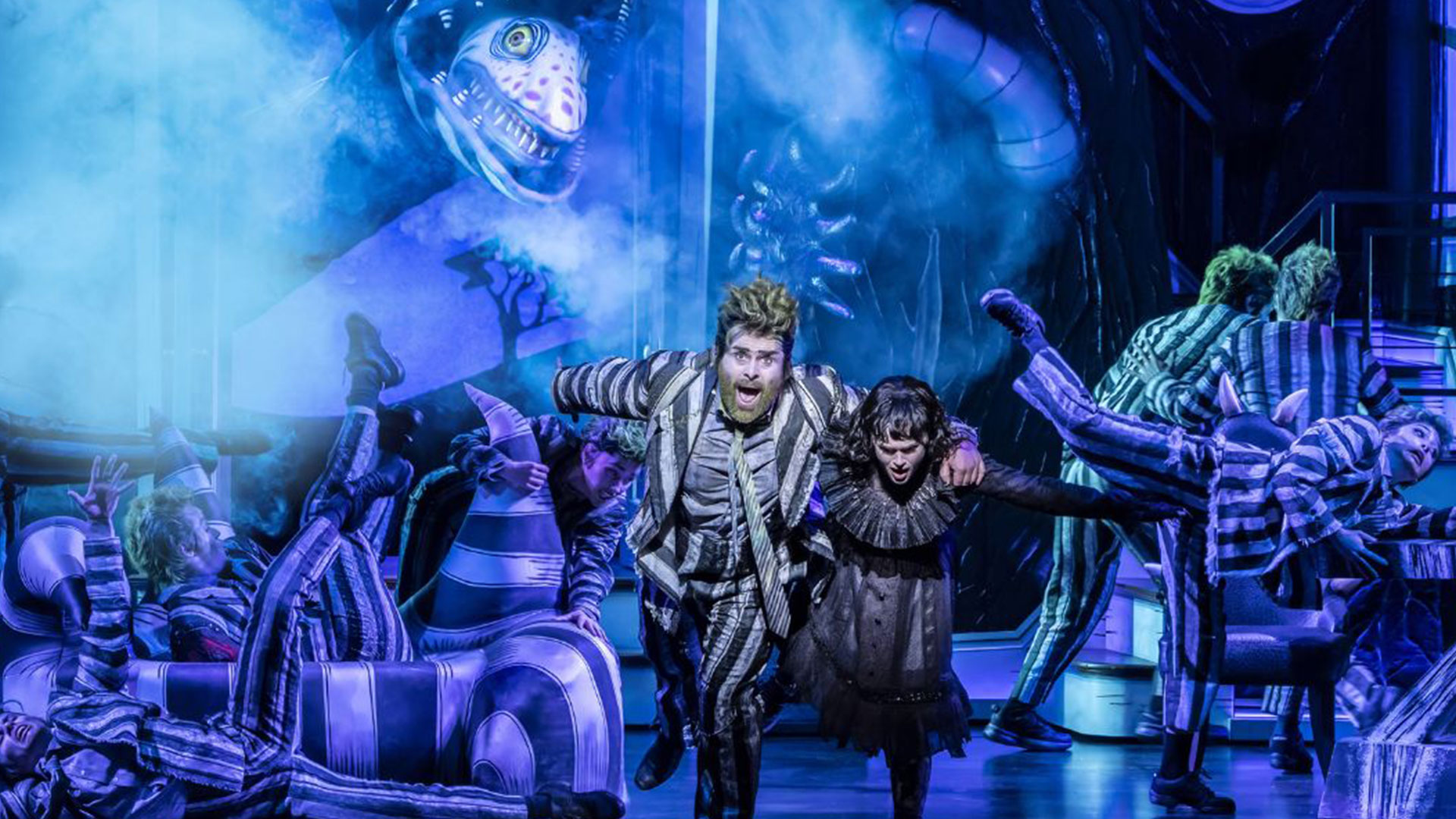 The two actors playing in Beetlejuice — one wearing the iconic stripy suit, the other a black lace dress — run onto the stage, fleeing from a giant snake.