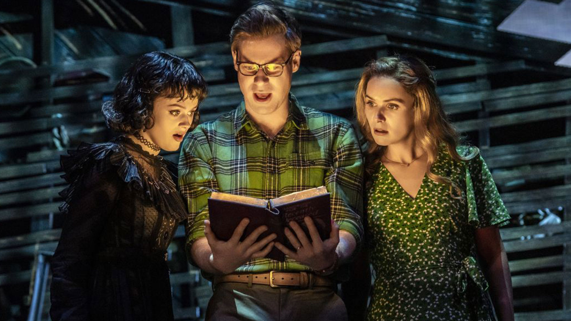 Immerse yourself in the world of Lydia Deetz in Beetlejuice the Musical, as she befriends a couple of ghosts and summons the spirit of Beetlejuice from the underworld. Image courtesy of London Theatre Direct. The actress playing Lydia Deetz in Beetlejuice stands on the left wearing a lacy black dress. She is standing next to the Maitlands, who are holding a book that is illuminating their faces as they read.