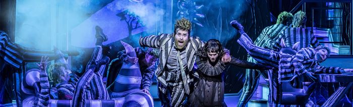 The two actors playing in Beetlejuice — one wearing the iconic stripy suit, the other a black lace dress — run onto the stage, fleeing from a giant snake.
