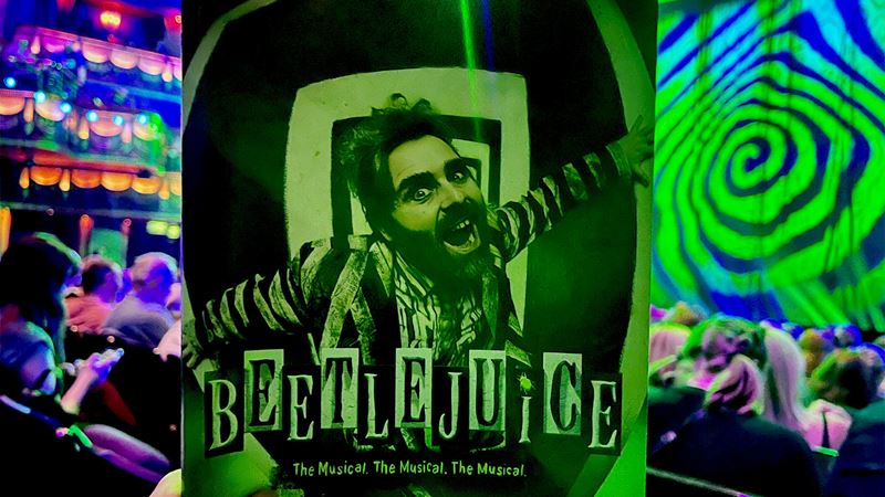 A programme for the musical Beetlejuice is being held by someone as they sit in the auditorium, with the stage lit up in green.