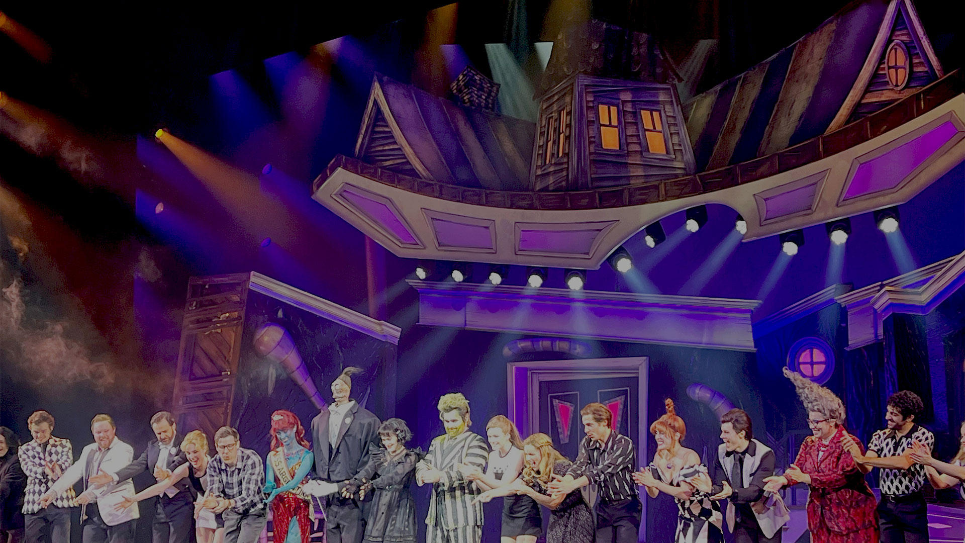 The full cast of Beetlejuice the Musical stands on stage, bowing to the applause of the audience. The Deetz house is lit up in purple and green in the background.