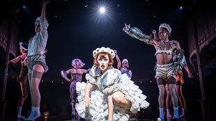 Katherine Langford wearing a white ruffled dress in the musical Cabaret at the Kit Kat Club, with dancers from the cast surrounding her on stage.