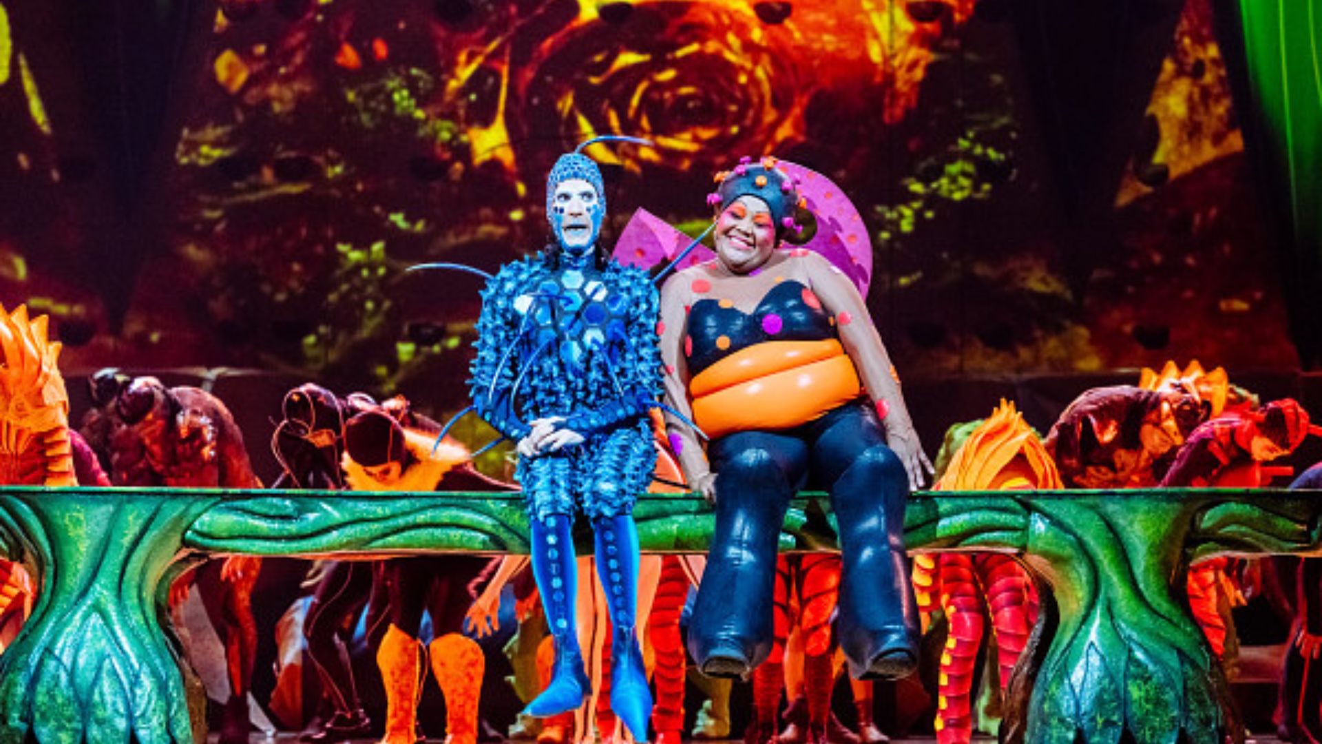 Two performers dressed as stylised insects sit on a leaf-like stage set during a Cirque du Soleil OVO performance, surrounded by vivid lighting.