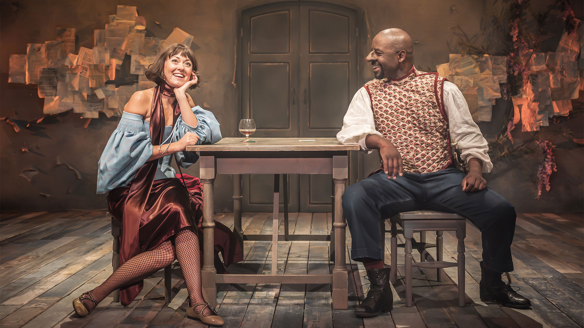 Adrian Lester, wearing dark jeans and a gold and red gilet, is sitting at a table looking at Susannah Fielding, who is sitting opposite him in a blue blouse and maroon silk skirt.