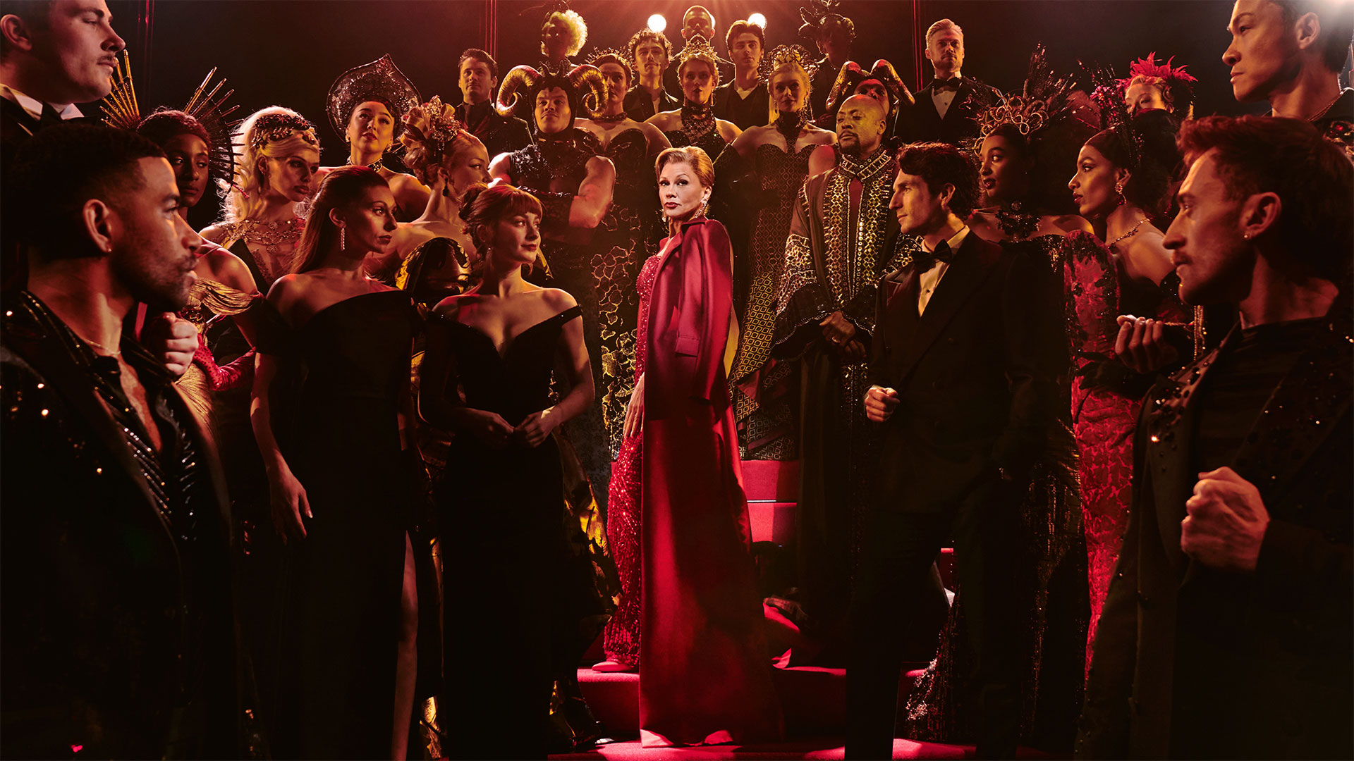 Miranda Priestly, played by Vanessa Williams, wears a glittering red dress and walks down the stairs surrounded by the cast of Devil Wears Prada.