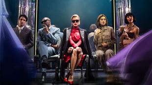 Actress Vanessa Williams, playing Miranda Priestley in the musical The Devil Wears Prada, is seated centre stage in a red dress and sunglasses, staring at the audience and surrounded by colleagues.