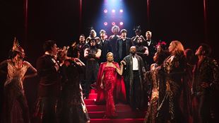 Miranda Priestly, interprétée par Vanessa Williams, porte une robe rouge scintillante et descend les marches entourée de la troupe du Diable s'habille en Prada.