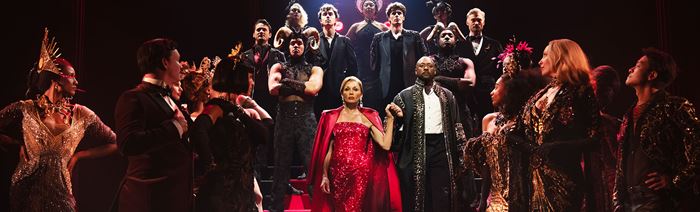 Miranda Priestly, interprétée par Vanessa Williams, porte une robe rouge scintillante et descend les marches entourée de la troupe du Diable s'habille en Prada.