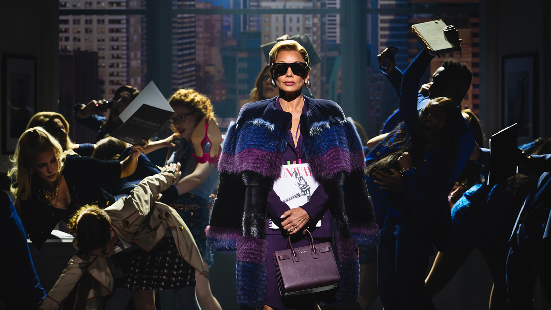 See London's latest musical sensation bring the iconic film The Devil Wears Prada to life on the stage of the Dominion Theatre. Image courtesy of Dewynters/ Photo credit: Matt Crockett. Vanessa Williams, portraying editor-in-chief Amanda Priesley, wears black sunglasses, a purple cape and a handbag as she walks across the stage while the press ask her questions.