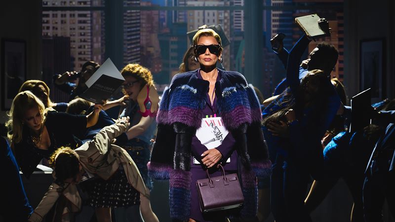 See London's latest musical sensation bring the iconic film The Devil Wears Prada to life on the stage of the Dominion Theatre. Image courtesy of Dewynters/ Photo credit: Matt Crockett. Vanessa Williams, portraying editor-in-chief Amanda Priesley, wears black sunglasses, a purple cape and a handbag as she walks across the stage while the press ask her questions.