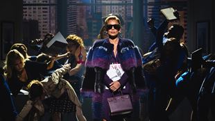 Vanessa Williams, qui incarne la rédactrice en chef Amanda Priesley, porte des lunettes de soleil noires, une cape violette et un sac à main. Elle traverse la scène pendant que la presse lui pose des questions.