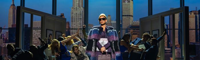 Vanessa Williams, portraying editor-in-chief Amanda Priesley, wears black sunglasses, a purple cape and a handbag as she walks across the stage while the press ask her questions.