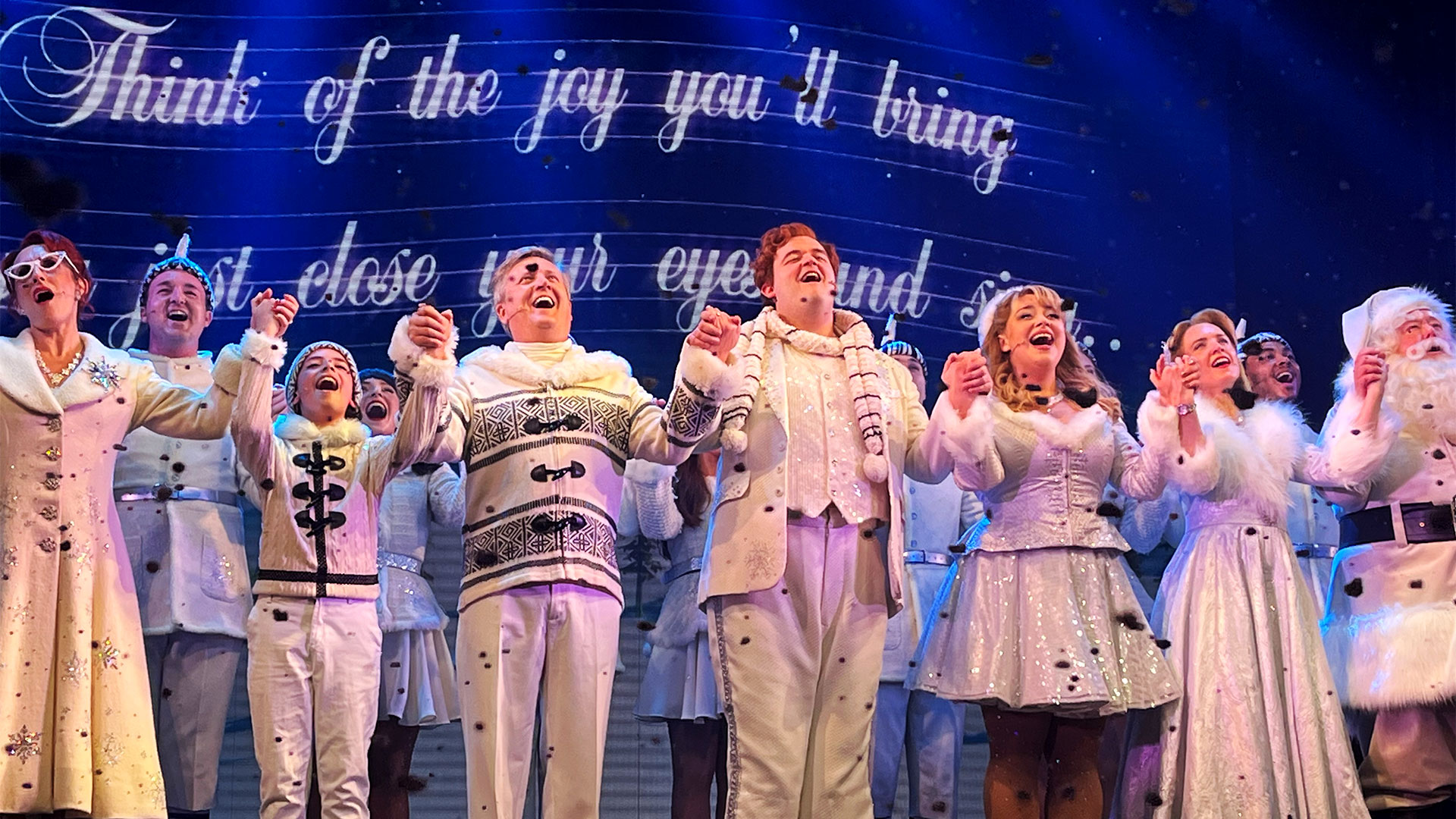 The cast of Elf the Musical, including Joel Montague and Carrie Hope-Fletcher, are all dressed in white and holding hands as they sing on stage together.