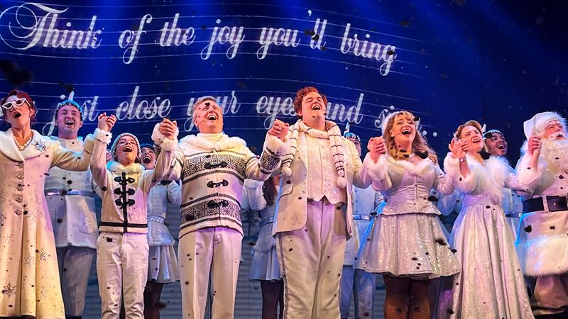 The cast of Elf the Musical, including Joel Montague and Carrie Hope-Fletcher, are all dressed in white and holding hands as they sing on stage together.
