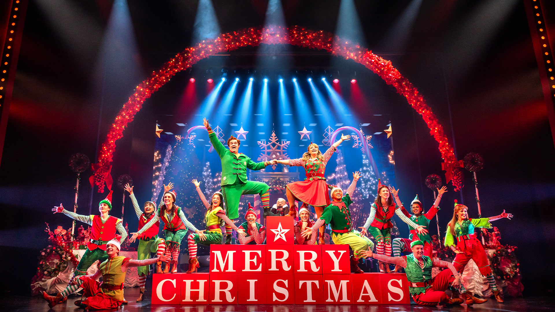 The cast of ELF The Musical is on stage, all dressed as elves on a stage decorated with giant candy canes and giant red cubes spelling 'Mery Christmas'.