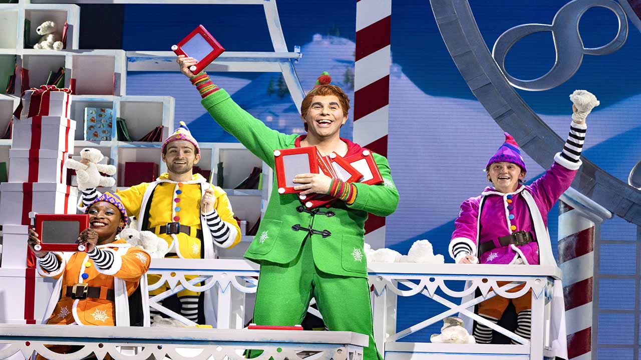 Der Schauspieler, der Buddy spielt, trägt eine auffällige grüne Uniform und hält ein paar Spielsachen in der Hand, umgeben von den kleinen Helfern des Weihnachtsmanns, die alle mit bunten Kostümen gekleidet sind.