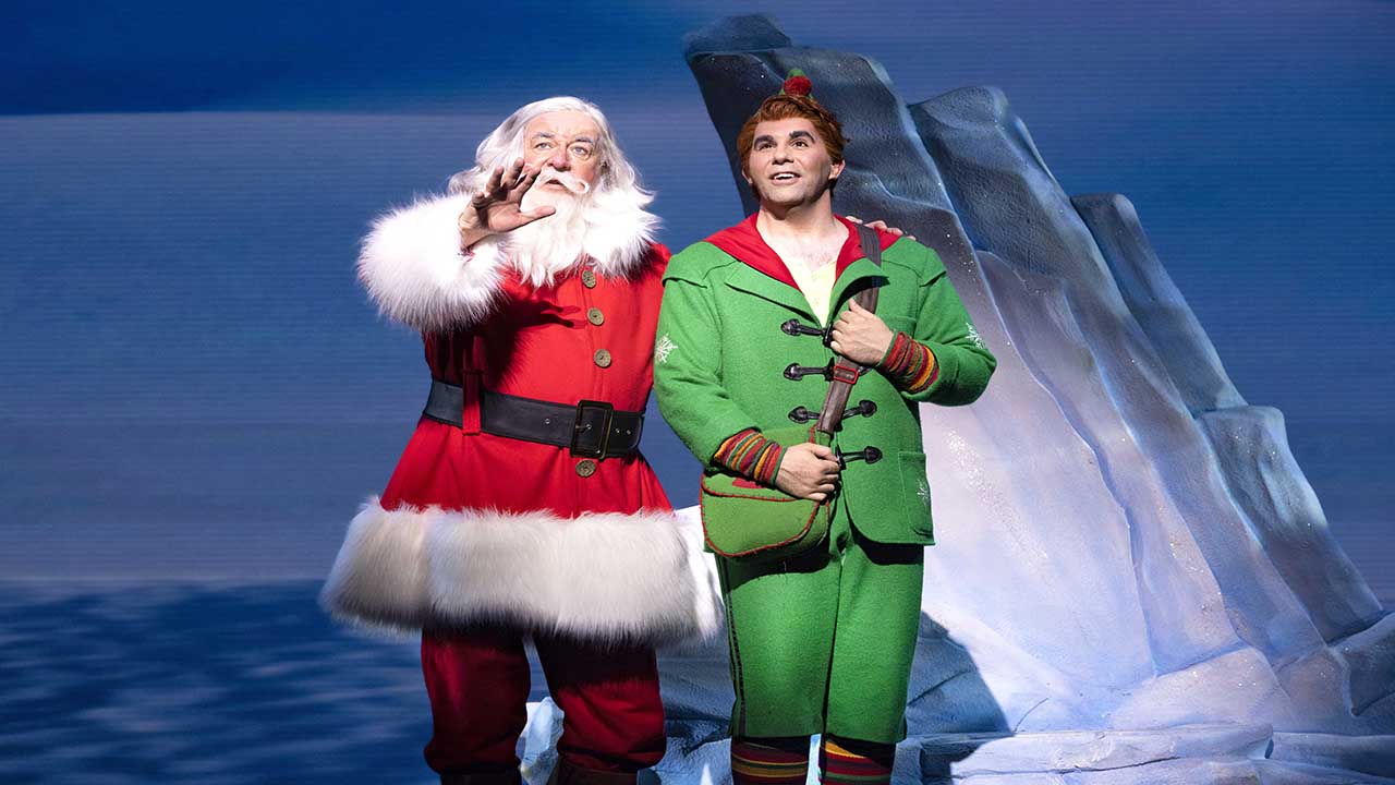 Erleben Sie Buddy auf der Suche nach der wahren Bedeutung von Weihnachten in ELF the Musical im Dominion Theatre. Bild mit Genehmigung von Dewynters Buddy in seinem tiefgrünen Elfenkostüm steht neben dem Weihnachtsmann auf der Bühne des London Dominion.