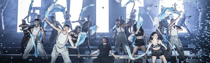 tiered-stage with actors and actresses cheering and waving flags, confetti falling down and bold light-up letters at the back of the stage spelling EVITA