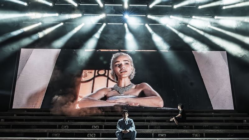 large tiered west end stage with an actor sat at the bottom of the tiered steps, and another actor sat a few rows behind looking up at a screen with a woman performing a song