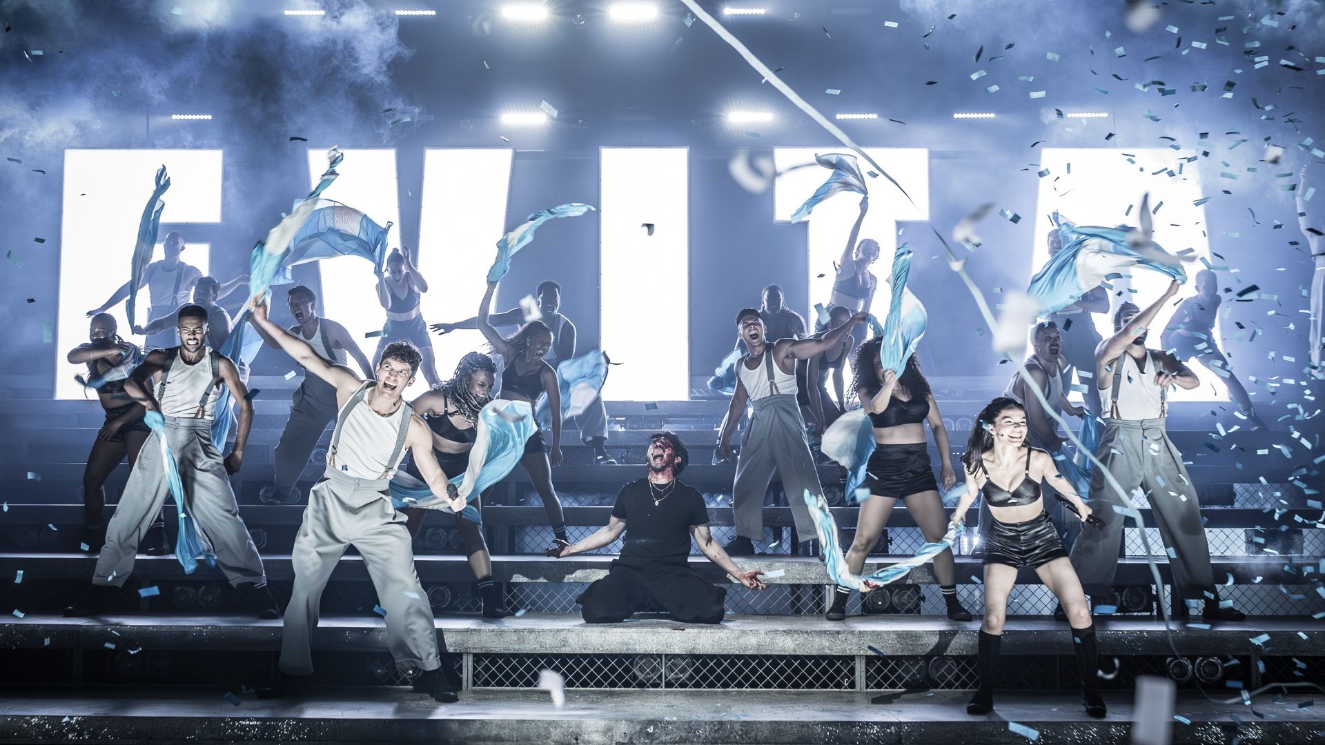 The revival of Andrew Lloyd Webber and Tim Rice's legendary musical Evita, starring Rachel Zegler, at the London Coliseum. Image courtesy of London Theatre Direct. tiered-stage with actors and actresses cheering and waving flags, confetti falling down and bold light-up letters at the back of the stage spelling EVITA