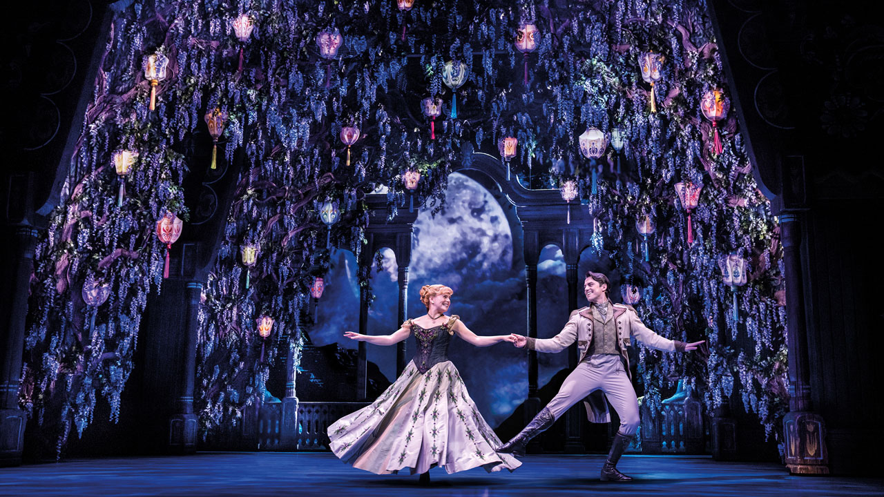 Les acteurs jouant Anna et Hans, vêtus de magnifiques tenues de bal, dansent dans une magnifique salle décorée de lanternes et de plantes, sous la pleine lune.
