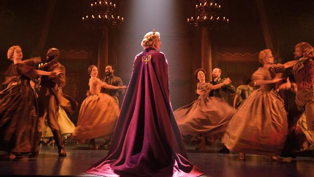 Die Schauspielerin, die Elsa im Musical Frozen spielt, trägt einen roten Umhang  auf einem Ball, auf dem Paare in Kleidern und dreiteiligen Anzügen um sie herum tanzen. 