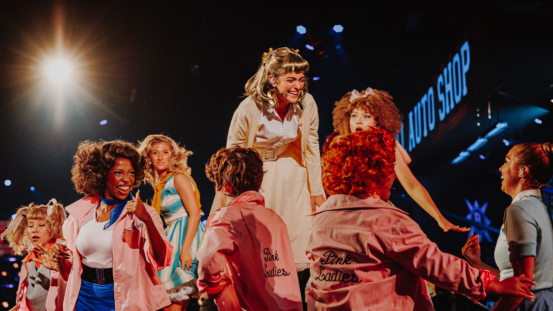 A group of actresses in pink jackets are smiling and talking to an actress in a white retro dress, who is portraying Sandy in Grease: The Immersive Experience.