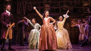 Three women, The Schuyler Sisters, stand together on stage with their arms in the air in large pastel dresses. 